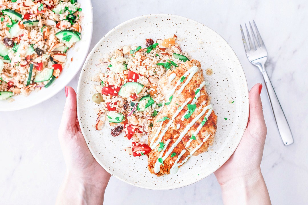 Hands holding plate of Moroccan-spiced rockfish on bed of couscous salad.