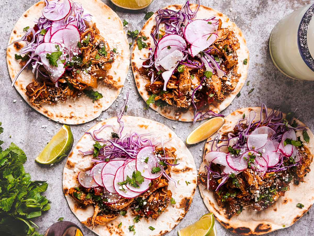 Several shredded chicken tacos on a gray background with lime wedges.