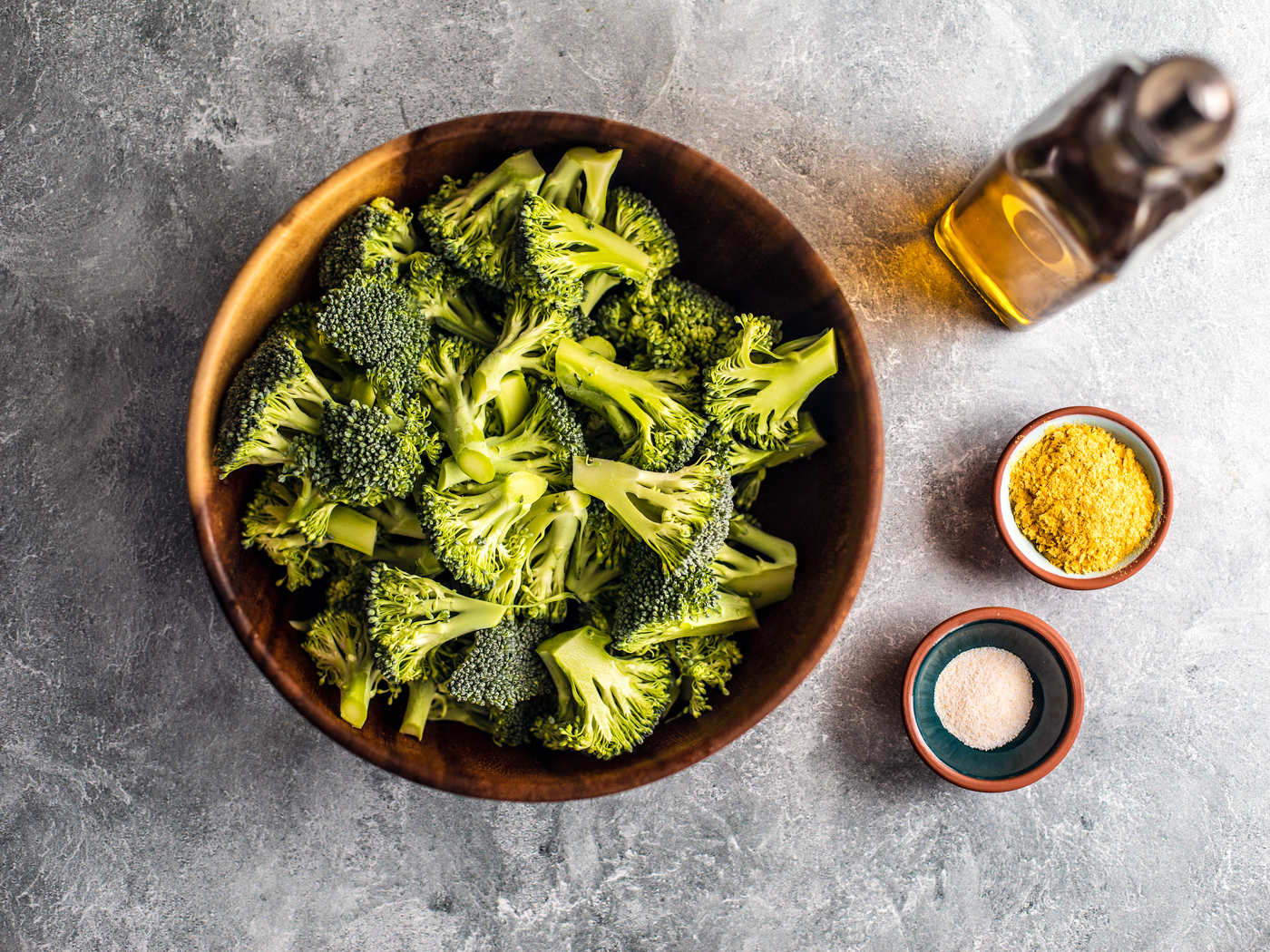 Bamboo bowl full of broccoli florets next to pinch bowls of garlic salt, nutritional yeast, and a bottle of olive oil.