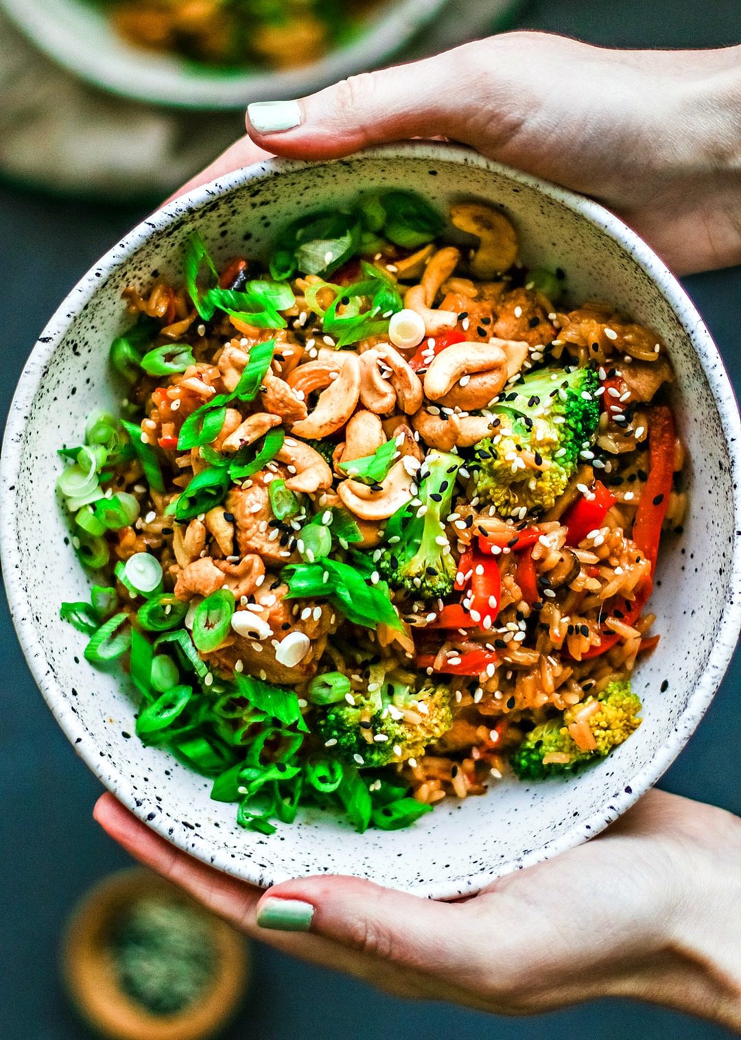 Hands holding bowl of Honey Garlic Chicken and Rice, loaded with veggies and cashews.