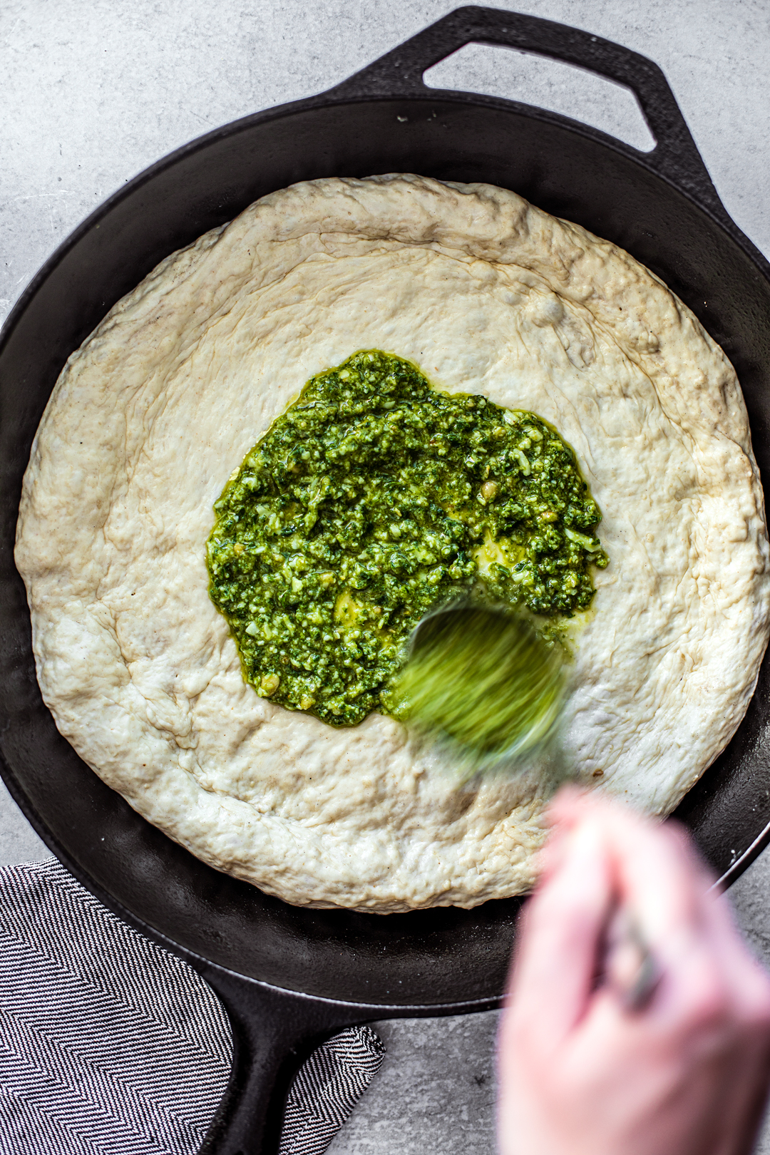 Pizza dough formed in skillet being topped with fresh pesto.