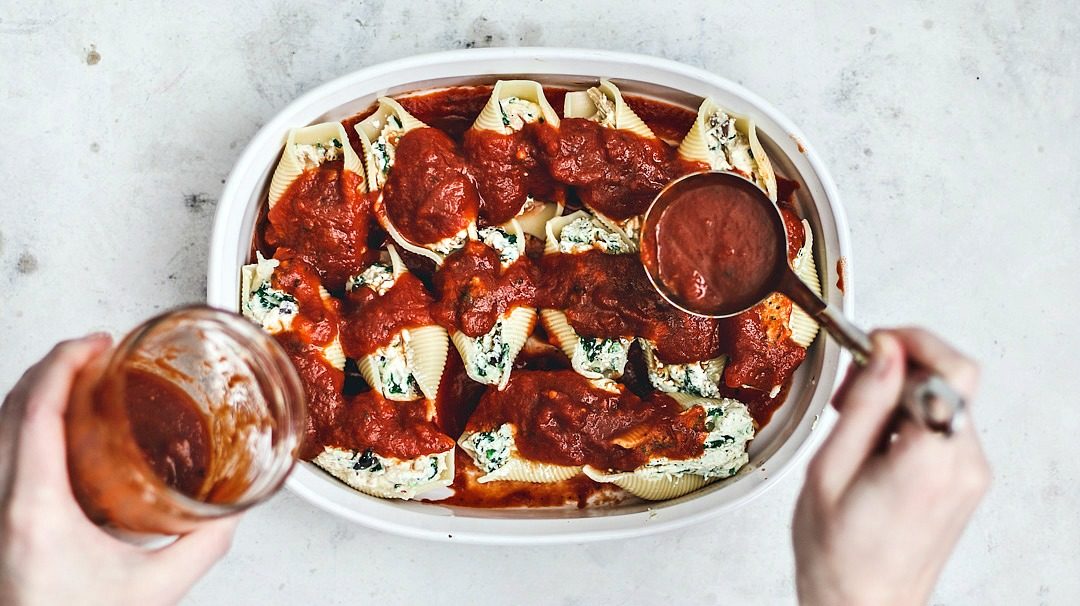 Sauce being spooned over stuffed shells in baking dish.