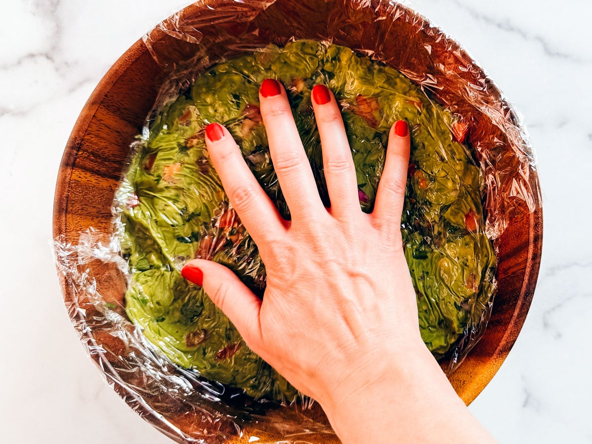 Hand pressing down on plastic wrap to cover guacamole.