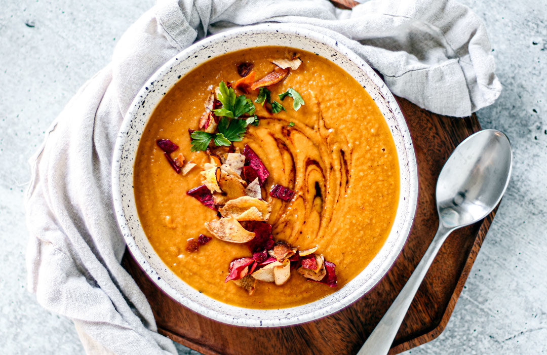 Bowl of sweet potato soup with colorful root chips and green herbs scattered over the top.