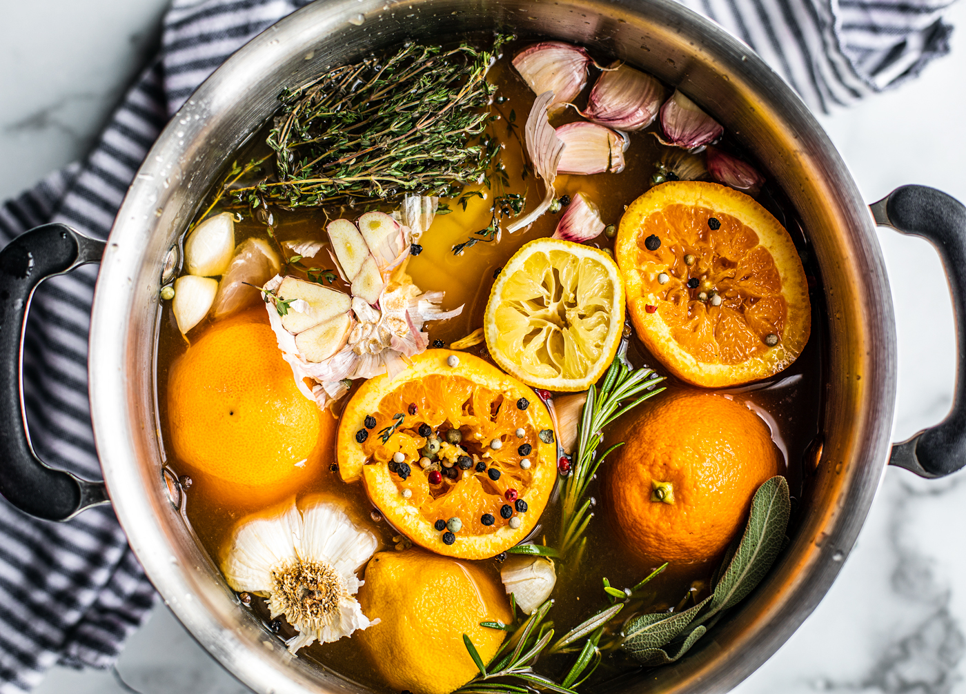 Large stock pot full of ingredients for turkey brine recipe: fresh herbs, oranges, lemons, peppercorns, and garlic.