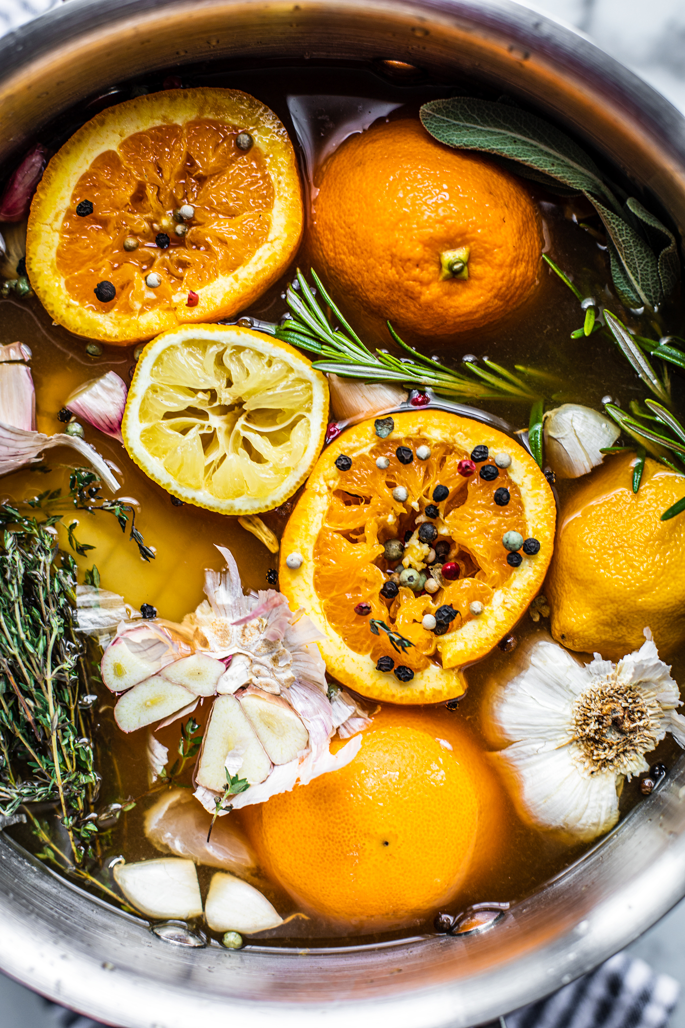 Large stock pot full of fresh herbs, oranges, lemons, peppercorns, and garlic.