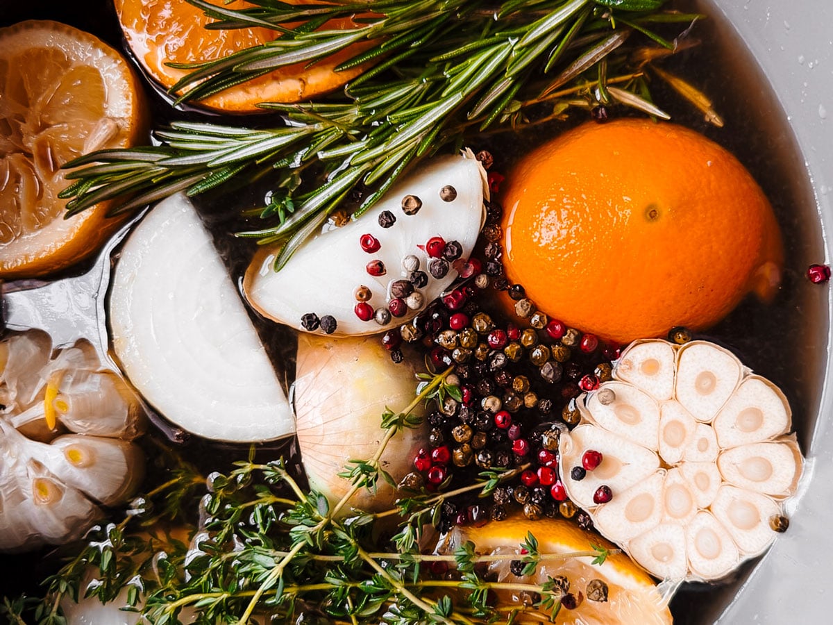 Citrus fruit, garlic, onions, and herbs floating in apple cider in a large pot.