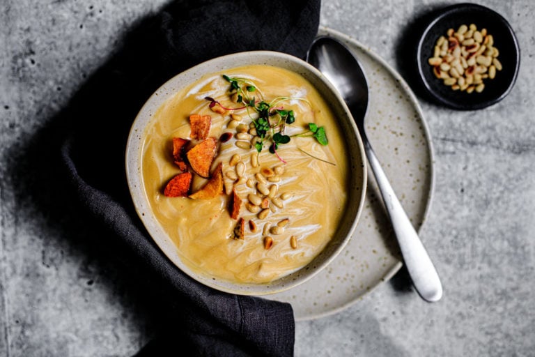 Bowl of roasted garlic and parsnip soup garnished with toasted pine nuts, root chips, and microgreens.
