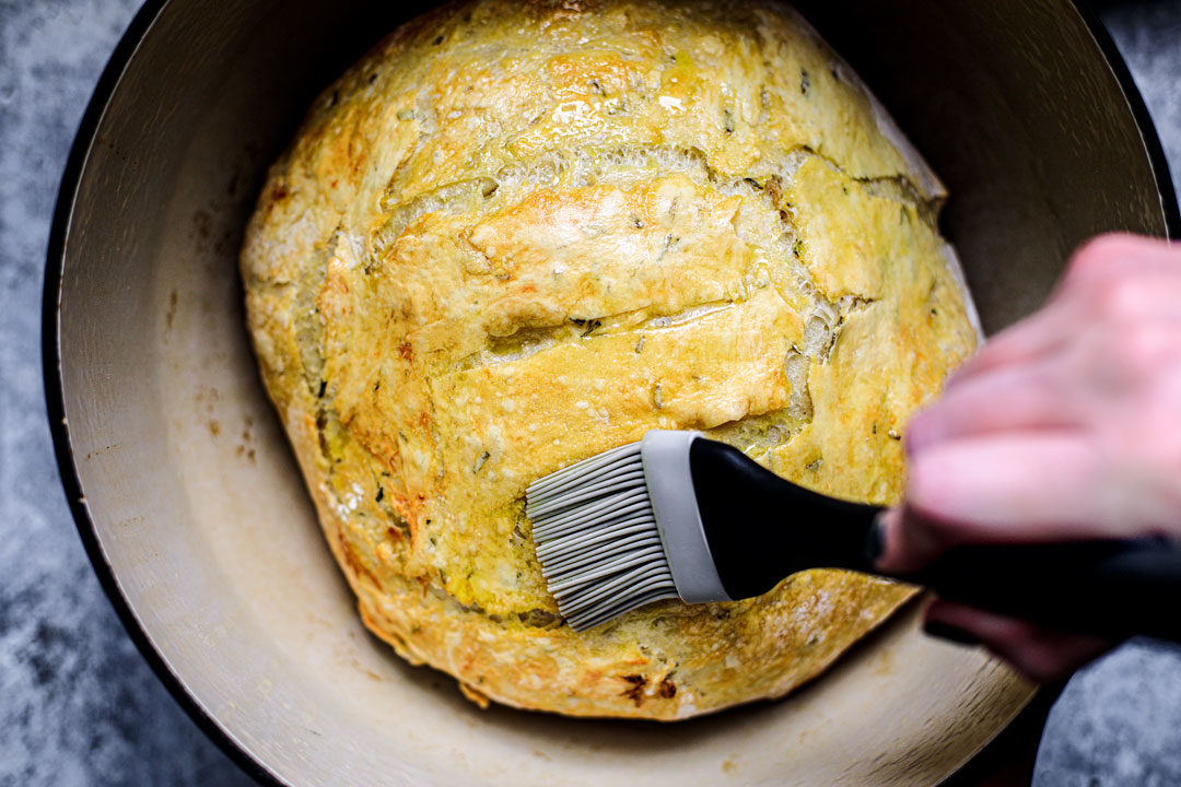 Baked bread in Dutch oven being brushed with olive oil.