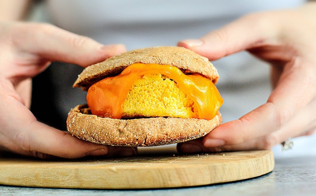 Hands holding cheesy egg muffin between English muffin bread.