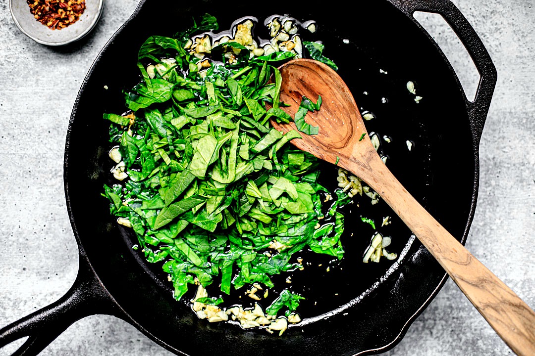 Skillet with oil, garlic, and spinach in it, with a wooden spoon.