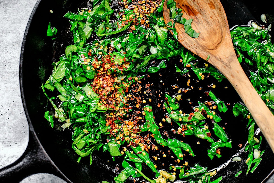 Skillet with cooked spinach and garlic, and the addition of dried chile flakes, with a wooden spoon.
