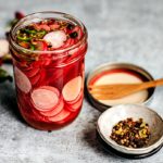 Side shot of jar of quick pickled radishes with pinch bowl of pickling spices.