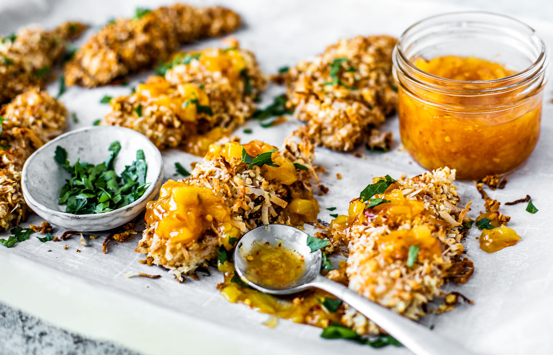 Side shot of crispy baked coconut chicken on a sheet pan with glass jar of spicy pineapple sauce.