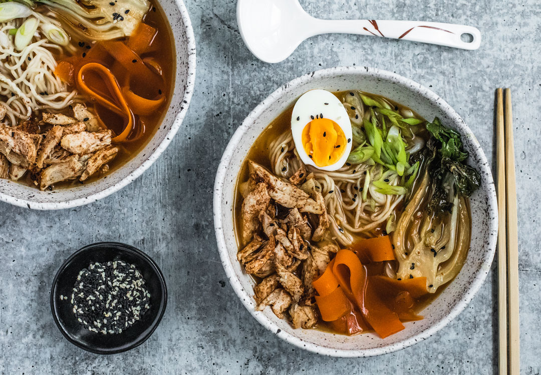 Two bowls of Five Spice chicken Ramen side by side, both topped with soft-boiled eggs, seared bok choy, and ribboned carrots.