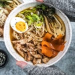Hands holding up five spice chicken Ramen bowl topped with an egg and vegetables.