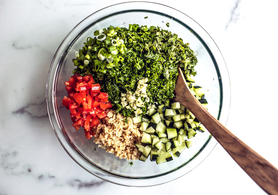 Chopped tabbouleh ingredients in a glass mixing bowl with a wooden spoon.