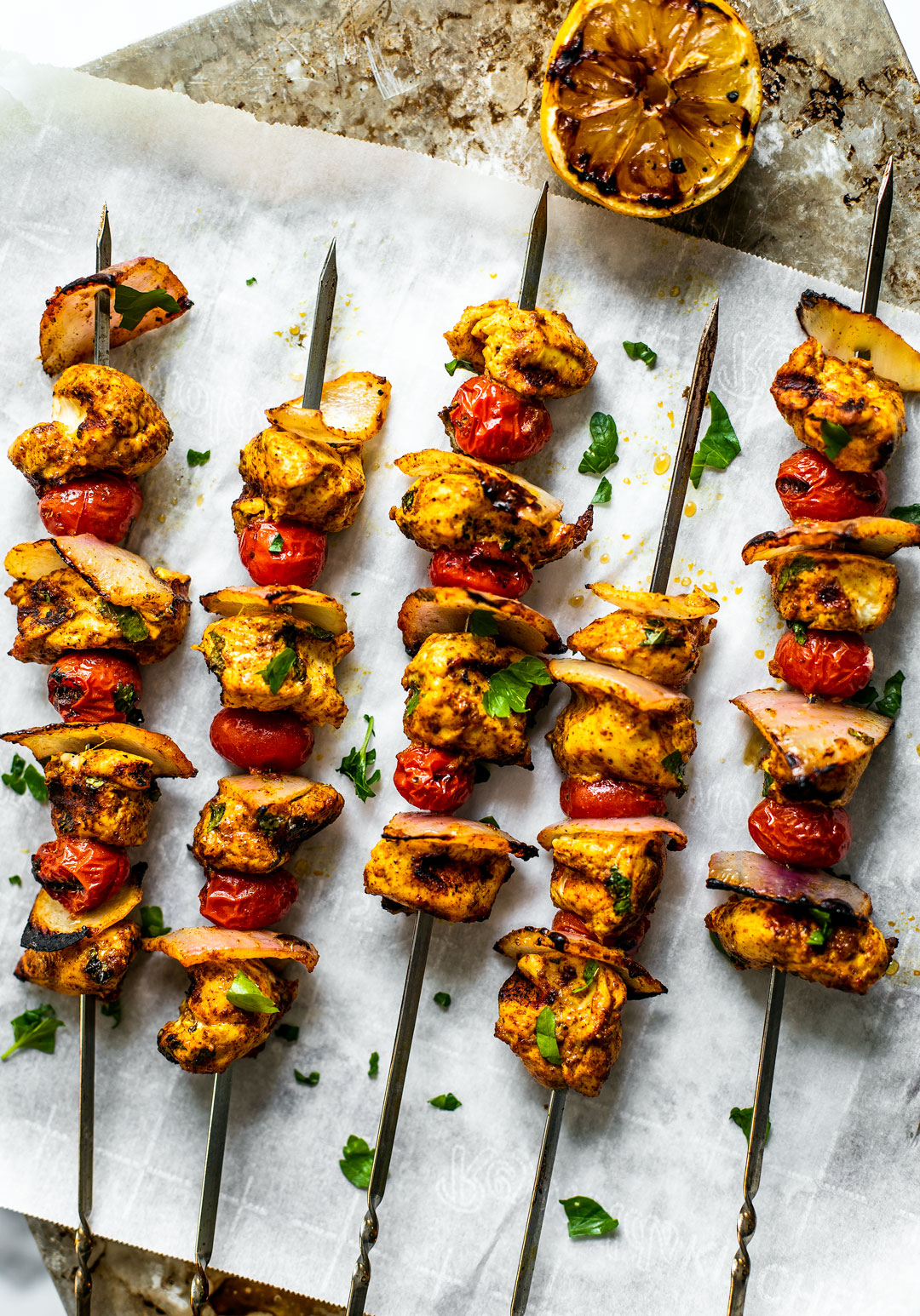 Grilled Moroccan chicken skewers set out over parchment paper with a grilled lemon half on the side.