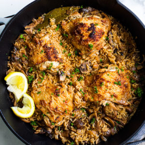 Close up of pan full of cooked rice, mushrooms, golden crisp chicken thighs, and lemon wedges.