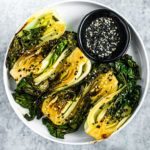 Round white plate stacked with bright green seared baby bok choy with a black pinch bowl full of black and white sesame seeds.