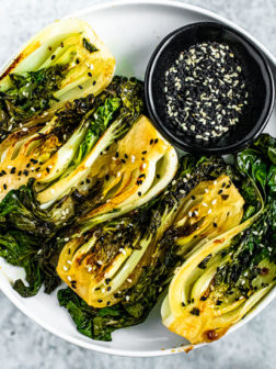 Round white plate stacked with bright green seared baby bok choy with a black pinch bowl full of black and white sesame seeds.