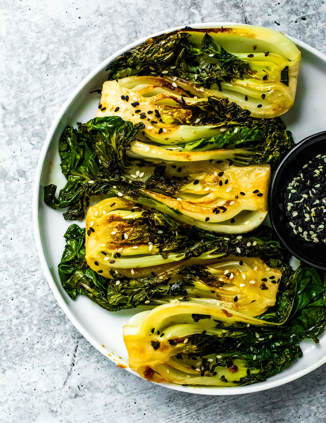 Close up of round white plate stacked with bright green seared baby bok choy with a black pinch bowl full of black and white sesame seeds.