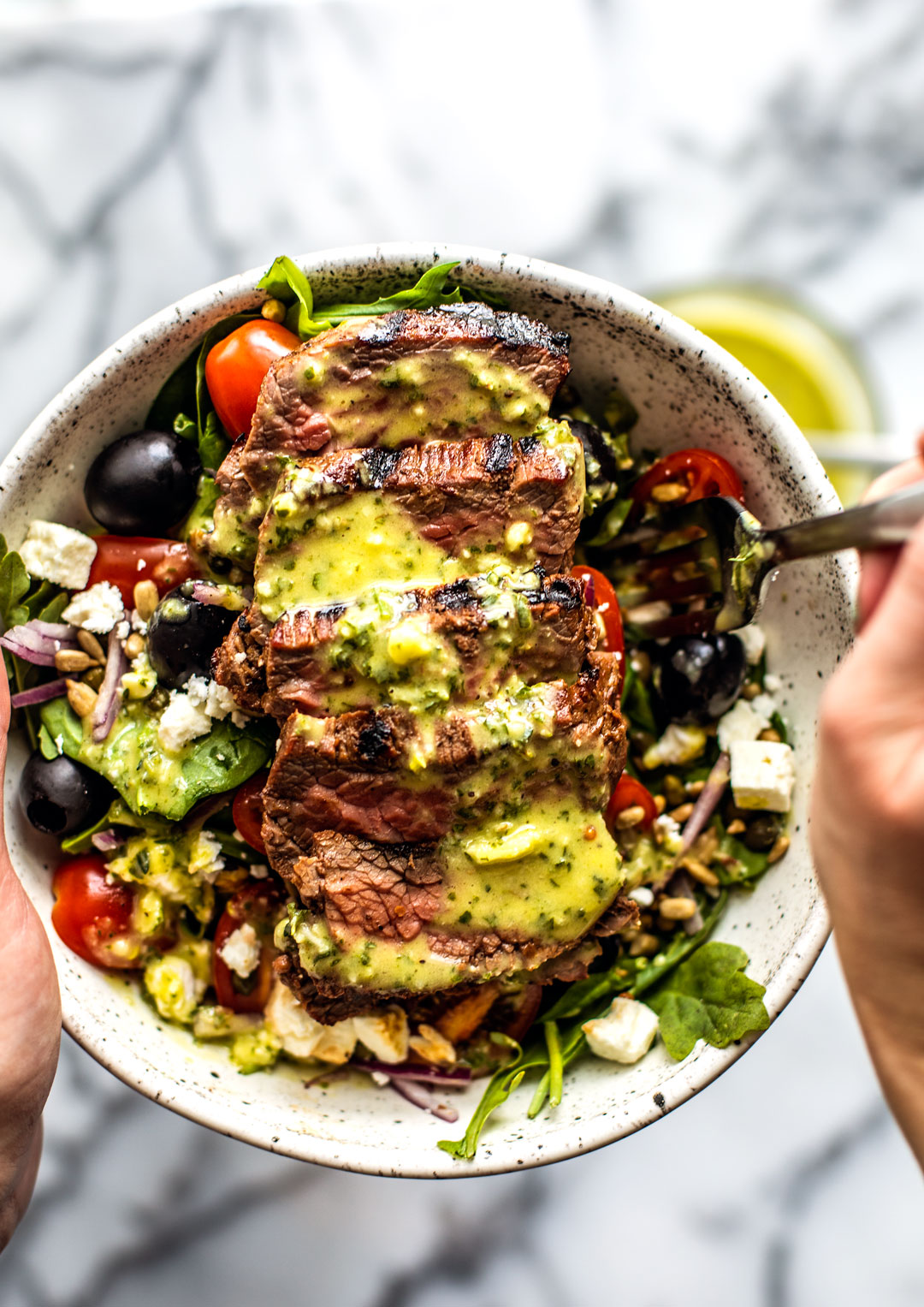 Hands holding up a bowl of Greek Steak Salad drizzled with herb and garlic vinaigrette.