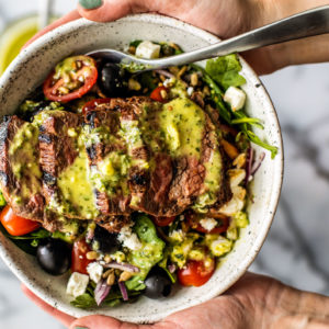 Hands holding up a bowl of Greek Steak Salad drizzled with herb and garlic vinaigrette.