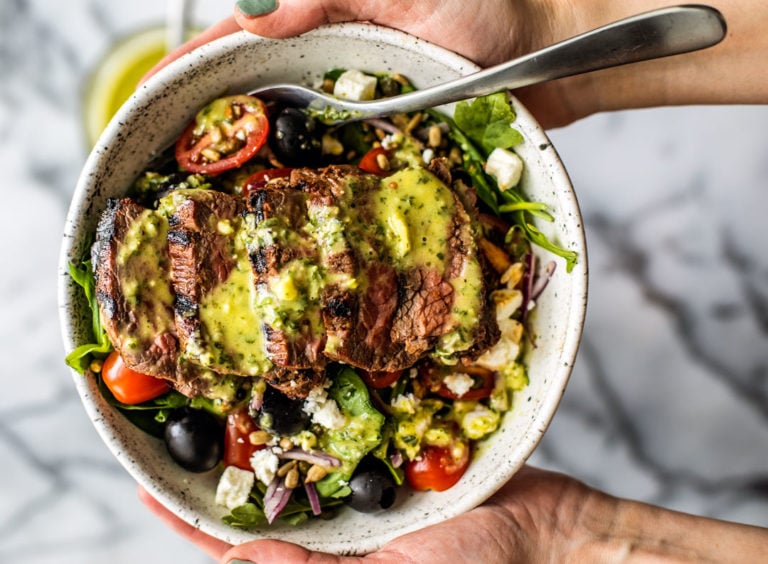Hands holding up a bowl of Greek Steak Salad drizzled with herb and garlic vinaigrette.