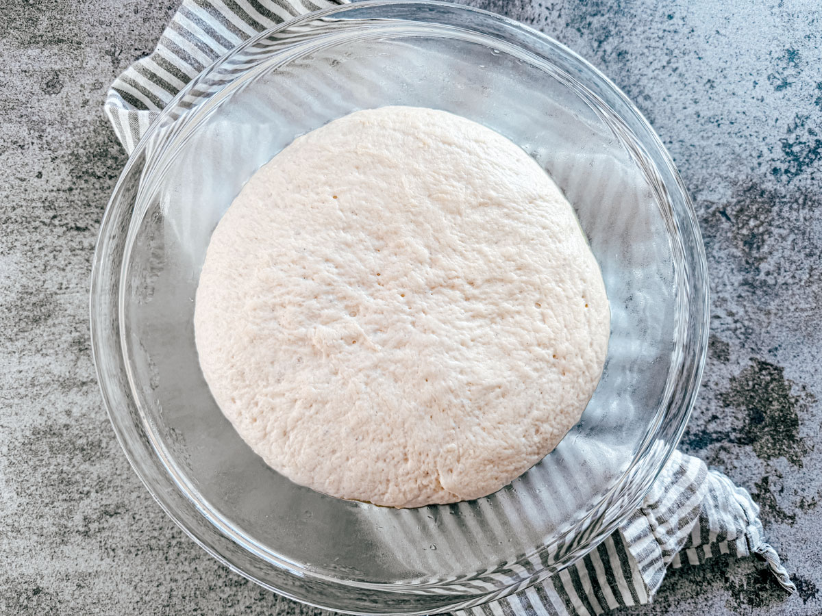 Risen pizza dough in a glass mixing bowl.