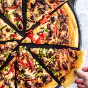 Sausage and roasted red pepper pizza slices on a cast iron pizza pan; a hand is reaching for one slice.
