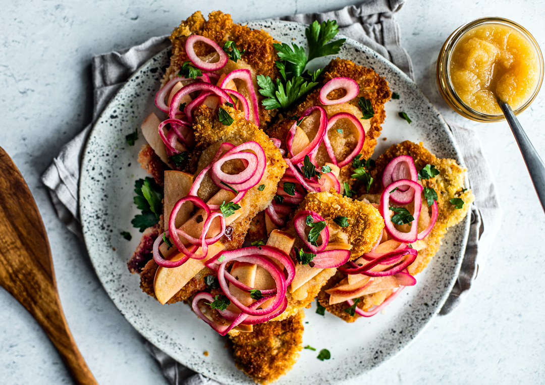 Serving plate full of crispy pork schnitzel topped with pickled onions and apples, and parsley.