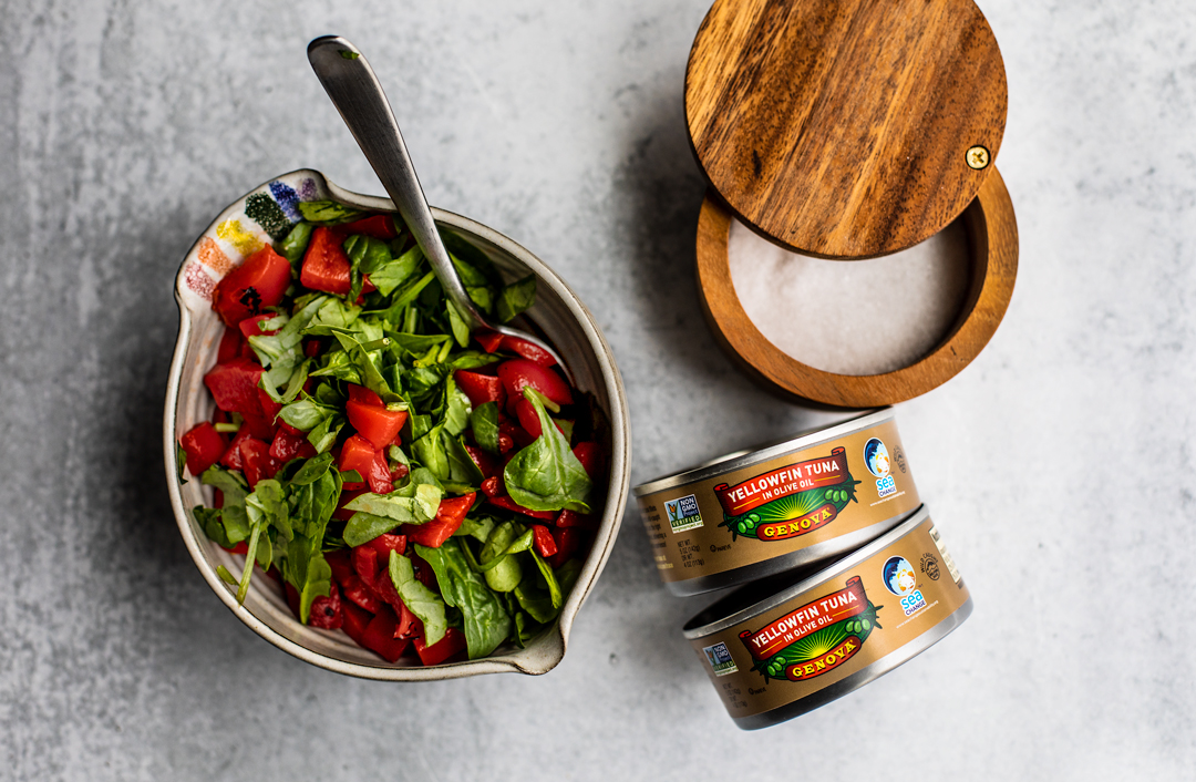 Bowl of spinach and roasted red peppers next to a salt crock and two cans of Genova tuna.