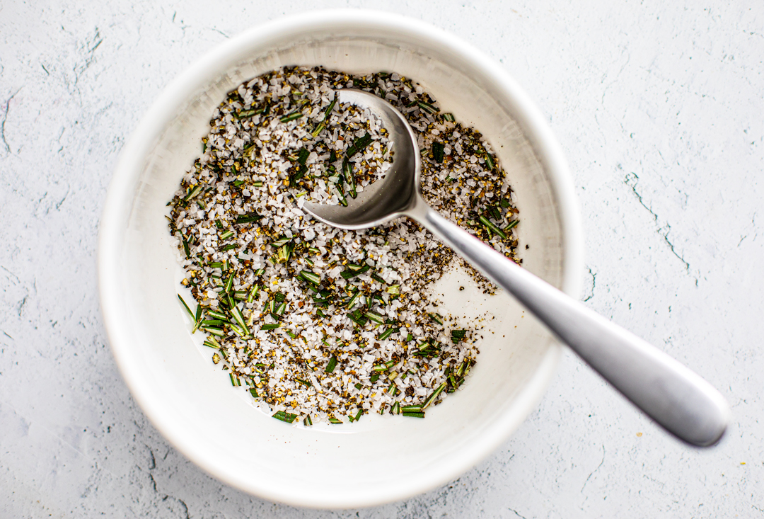 Small white bowl containing dry rub mix of coarse kosher salt, cracked pepper, and fresh rosemary.