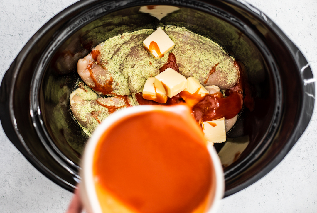 Slow cooker full of uncooked chicken, seasoning, and butter, with a hand pouring hot sauce over it.