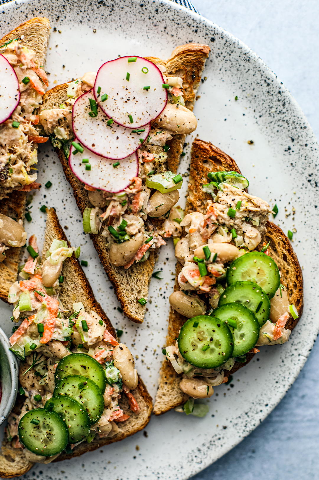 Overhead close up of fancy toasts topped with mackerel and white bean salad, radishes, and cucumbers.