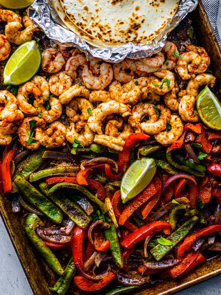Sheet pan covered with peppers, onions and shrimp with some lime wedges and soft shell tortillas on the side.