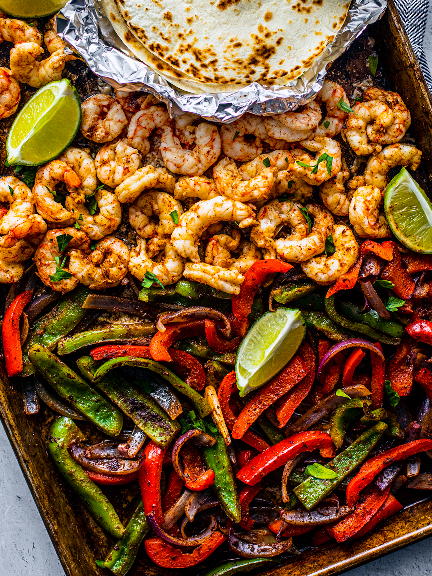 Sheet pan covered with peppers, onions and shrimp with some lime wedges and soft shell tortillas on the side.