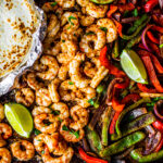 Sheet pan covered with peppers, onions and shrimp with some lime wedges and soft shell tortillas on the side.