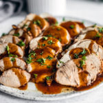 Side shot close up of platter of sliced pork tenderloin drizzled with gravy and garnished with parsley.