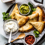 Overhead shot of platter of baked quesadillas with guacamole, salsa, and sour cream.