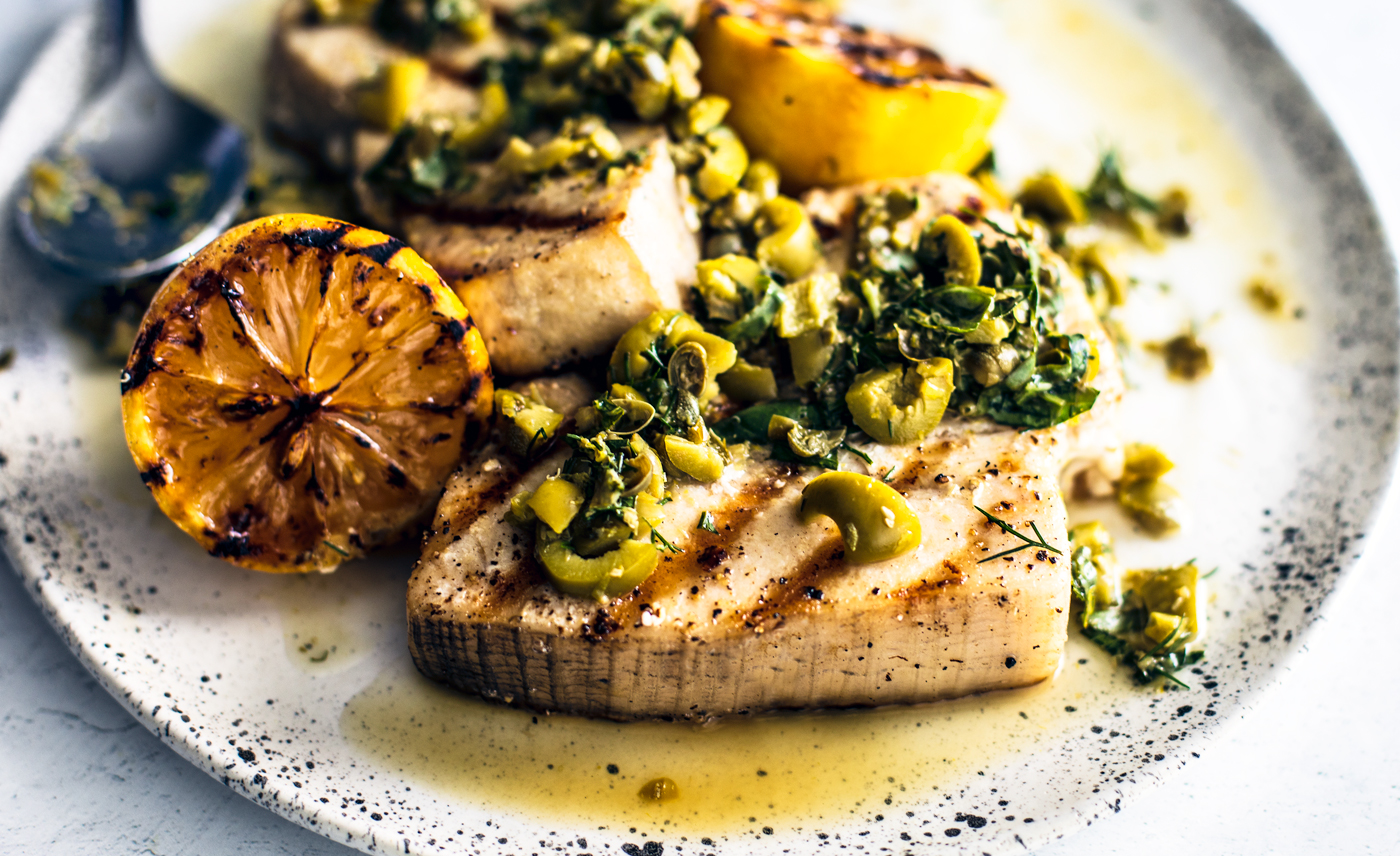 Close up side shot of swordfish steak with olives and herbs on top.