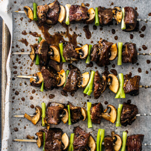 Overhead shot of skewers with beef, scallions, and mushrooms on parchment.