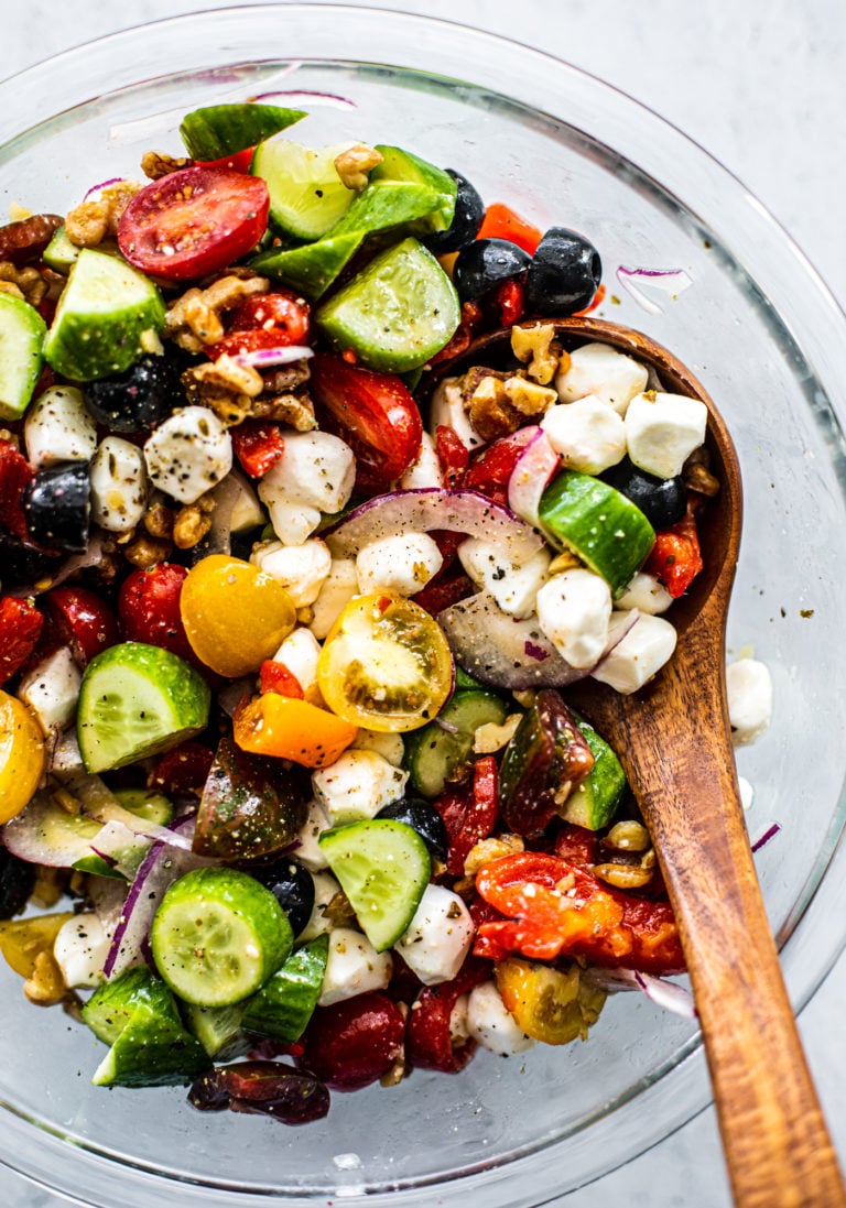 Glass serving bowl of Greek Cucumber Tomato Salad with a wooden serving spoon.