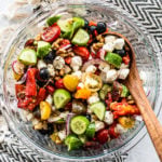 Glass serving bowl full of Greek cucumber salad with a wooden serving spoon in the bowl.