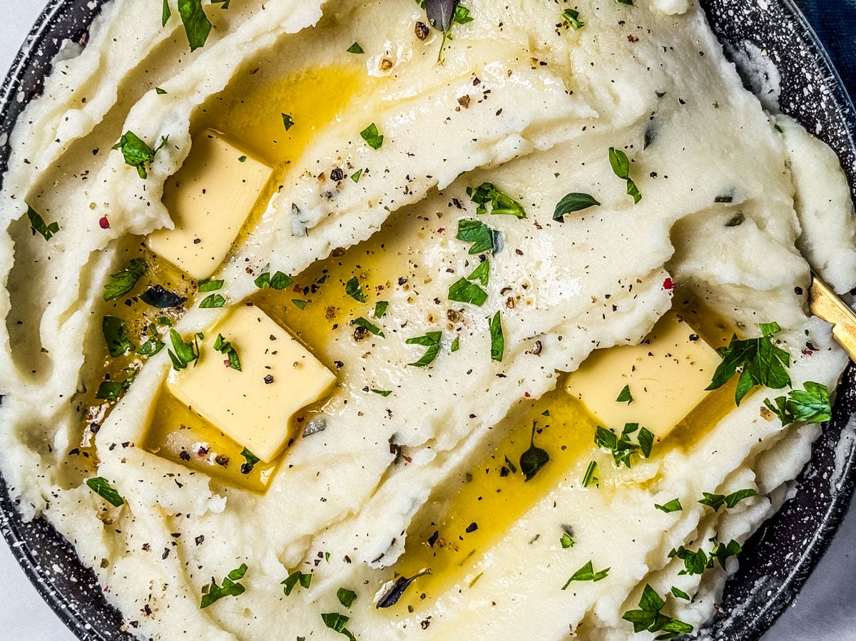Bowl of creamy mashed potatoes garnished with knobs of melting butter and herbs.