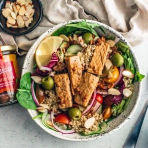 Big bowl full of mixed greens topped with quinoa, tomatoes, olives, cucumbers, almonds, and large chunks of tuna next to glass jar of King Oscar tuna.