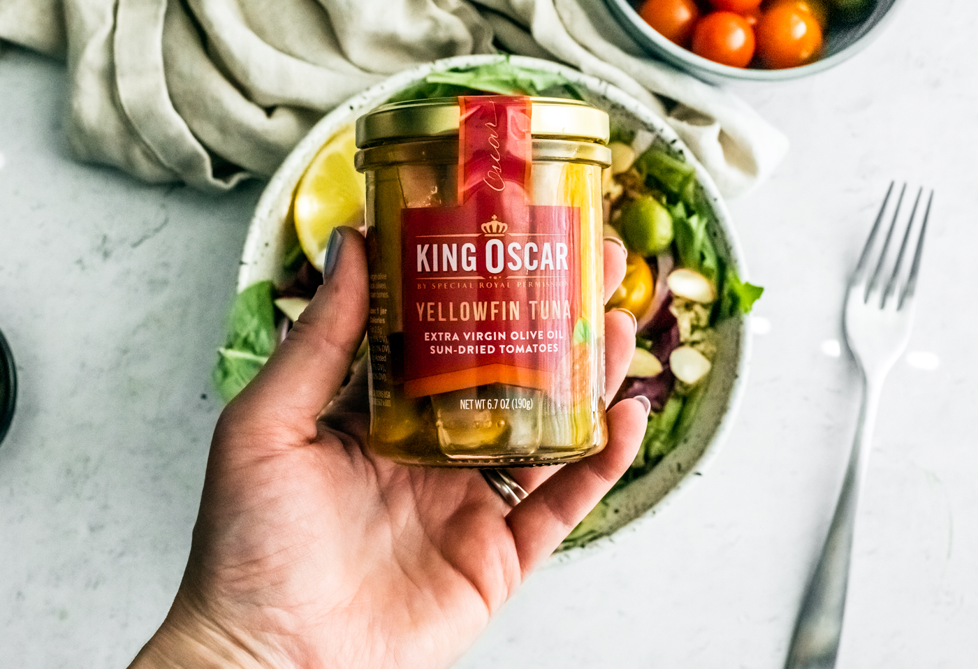 Hand holding up glass jar of King Oscar tuna fillets over salad in background.