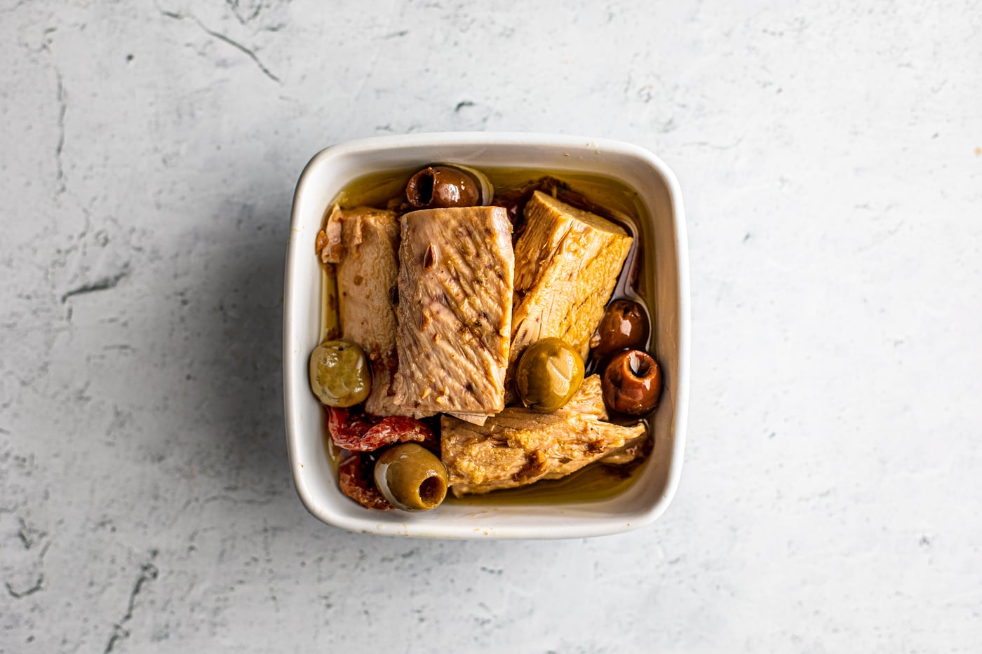 Overhead shot of big tuna fillets in a small serving dish with green and kalamata olives, oil, and sun-dried tomatoes.