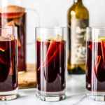 Three tall glasses of red sangria with sliced green apples and cinnamon sticks in it.